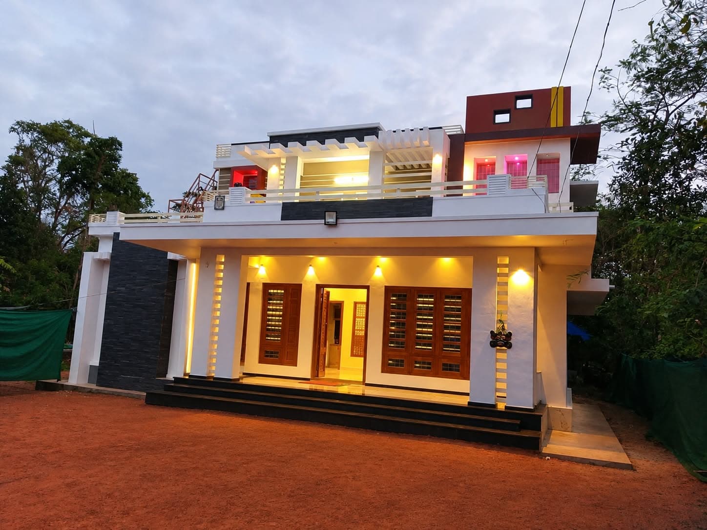Contemporary Kerala house facade at dusk with warm wall-washer lights, stone cladding and wooden windows, by Decorators India
