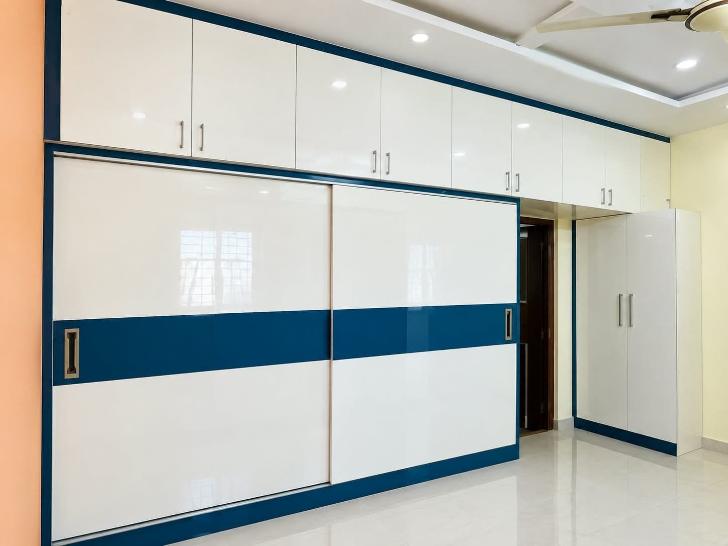White high-gloss sliding wardrobe with blue accent band and full-width loft cabinets